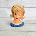 Front of blonde lady head vase with butterfly hair ornament, pearl-style necklace and blue ruffled neckline
