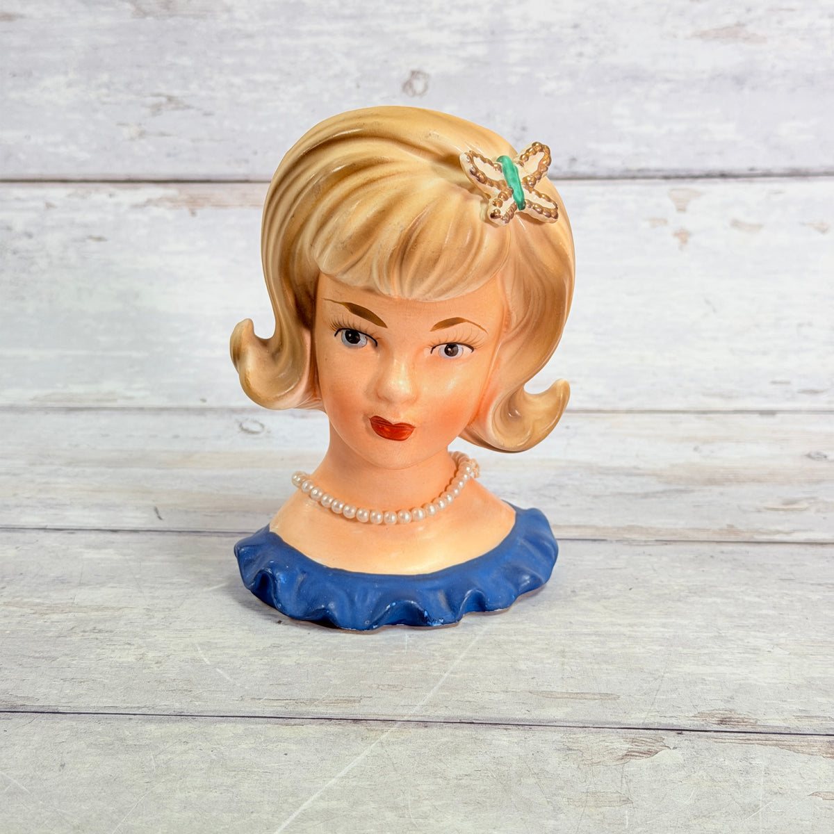 Front of blonde lady head vase with butterfly hair ornament, pearl-style necklace and blue ruffled neckline