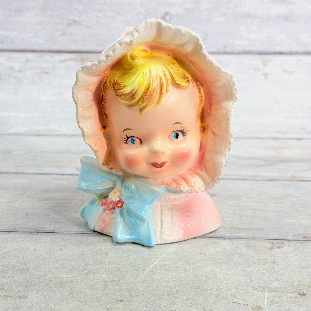 Front of child vase with pink bonnet, blue bow and flowers