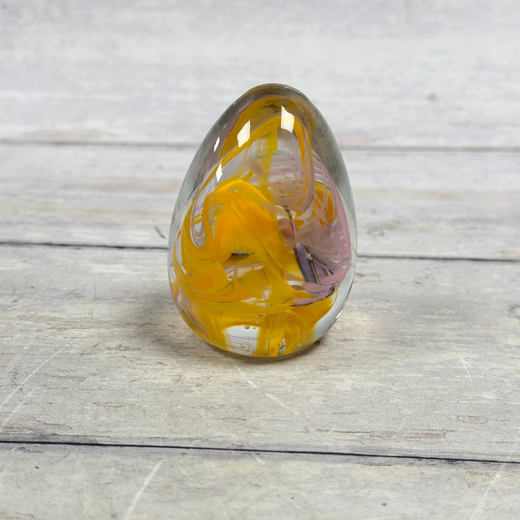 Clear glass egg paperweight with golden yellow and pale pink ribbon swirls