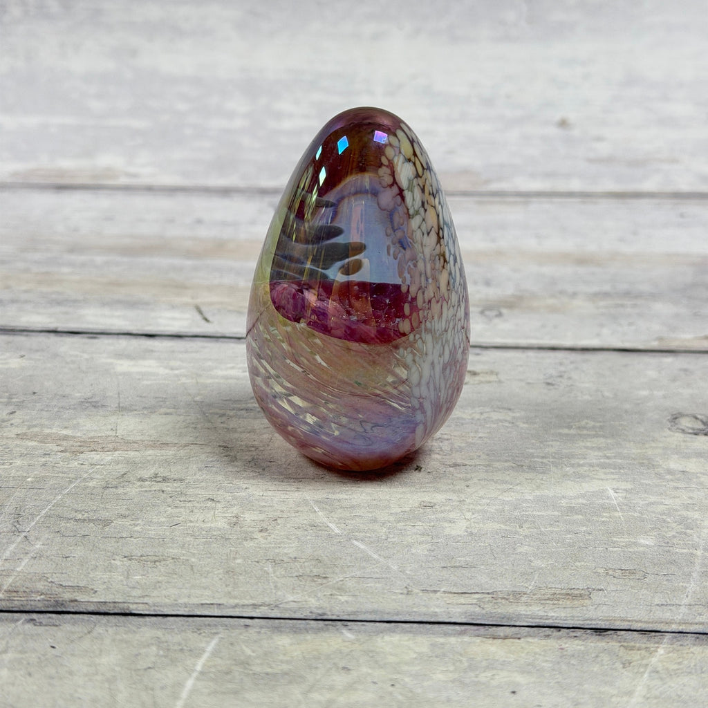 Side view of Glass Eye Studio pink and lavender iridescent egg paperweight with white feather-like decoration